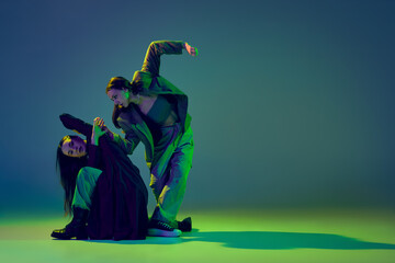 Relationship. Young stylish flexible girls in modern attires dancing contemp style dance isolated over gradient blue-green background at dance hall in neon light.