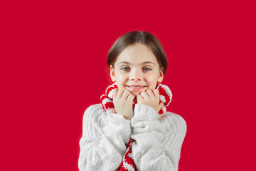 Closeup of a smiling face of young girl in a white sweater and striped scarf, standing isolated over red background wall looking at camera