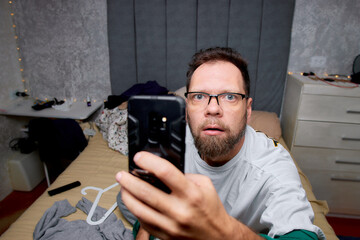 a bearded man in glasses with a smartphone on the bed is surprised by something looking at the phone display