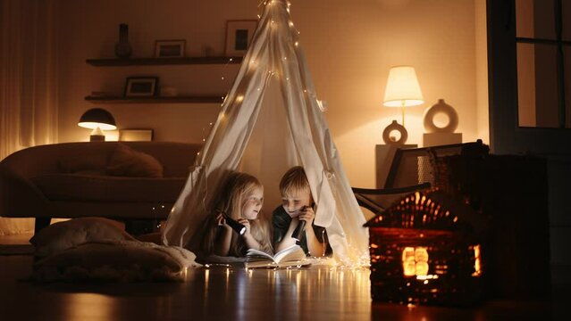 brother and sister children are playing in handmade teepee tent from blankets in home in darkness