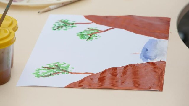 Kid draws a sea, trees  with watercolor paints and brushes, children's joint creativity. Child in the process of drawing. Close-up brush drawings with watercolor paint on white paper. 
