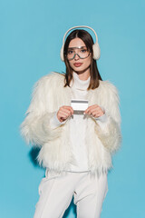 young woman in eyeglasses and stylish faux fur jacket holding credit card on blue.