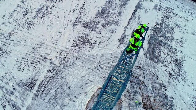 Cable Riggers Wearing Green Safety Suits And Hard Hats, Doing Cable Repair On Overhead Transmission Tower On A Winter Morning. - Drone POV