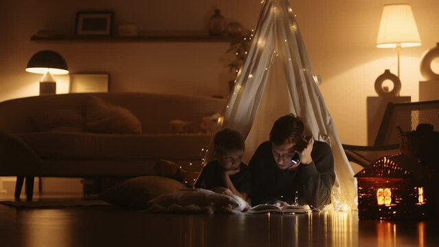 father is reading fairytales for preschooler son, man and boy in teepee in living room in evening