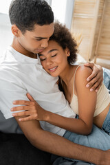 Fototapeta premium pretty and curly woman with closed eyes embracing with african american boyfriend in bedroom.