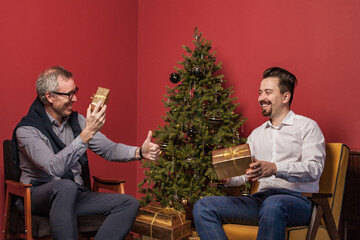 Elderly man and his son sitting on armchaiirs and exchanging Christmas presents against red studio background and Christmas tree