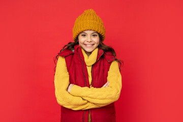 smiling child in knitted winter hat and down vest on red background, cold season
