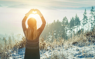 Happy woman stay on mountain top and making of love heart by hands in sunny frozen winter day.
