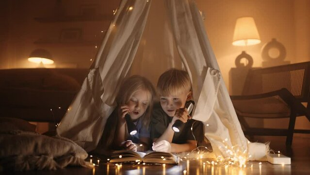 caucasian children inside tent from blanket in living room, cozy decorative lights in darkness