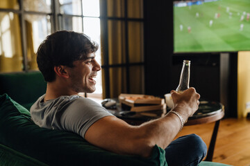 Young white man watching football match and drinking beer in front of the TV screen