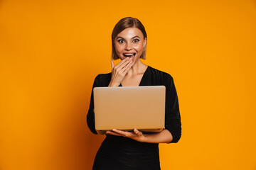 Surprised young woman using laptop covering her mouth isolated over orange background