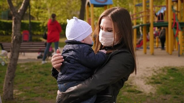 A young mother in a protective medical mask hugs her little one-year-old son in a Park against the backdrop of a crowded Playground. There's a coronavirus outbreak in the city.
