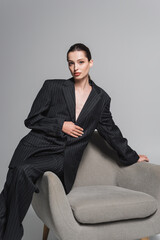 Fashionable woman in suit posing near armchair on grey background.