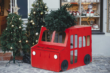 Stylish cardboard red car with christmas tree at festive building exterior. Modern simple christmas decor in city street. Winter holidays in Europe. Merry Christmas