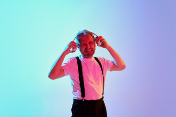 Portrait of mature man in shirt posing in headphones isolated over gradient purple background in neon light. Unpleasant sound