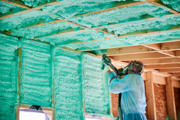 Male builder insulating wooden frame house. Man worker spraying polyurethane foam inside of future...