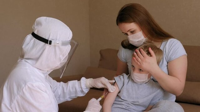A doctor in a protective suit and mask gives an injection to a little boy at home. Vaccination against coronavirus at home.