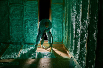 Male builder insulating wooden frame house. Man worker spraying polyurethane foam on floor inside...