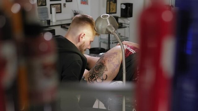 Portrait of a tattoo artist with a beard in a modern tattoo parlor, he holds a tattoo machine in his hand and stuffs a tattoo on a young woman's leg.
