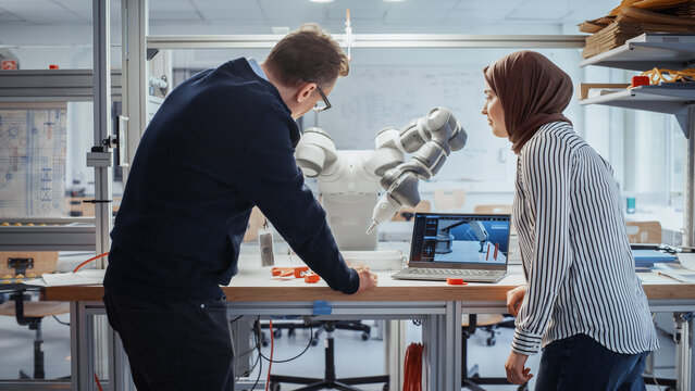Senior Male Engineer Explaining Controls to Muslim Female Computer Science Specialist and Pointing at Robotic Hand In the Research Laboratory with Laptop. Innovative Startup Company Concept.