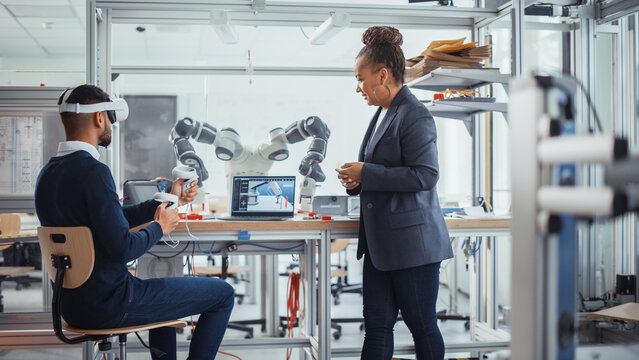 Black Senior Engineer Talks to Young Male Computer Scientist Who is Wearing Virtual Reality Headset and Uses Controllers to Remotely Manipulate Robotic Hand. High Tech Research Laboratory Concept - Powered by Adobe