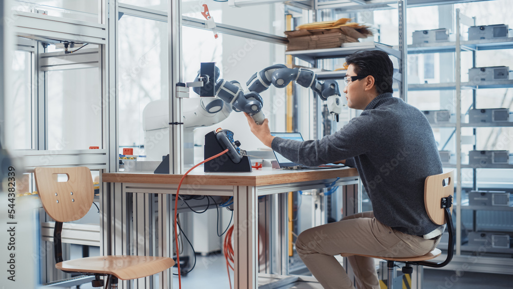 Leinwandbilder Asian Engineer Moving Robotic Arm with Control Panel While Developing Program on Laptop. Robotics Specialist Designs Plans and Processes Needed to Build Robots. Computer Science Laboratory.