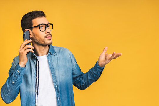 Oh, great news! Smiling young bearded casual man in glasses talking on the mobile phone isolated over yellow background.