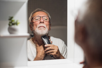 Senior man is trimming his beard with electric trimmer in bathroom.