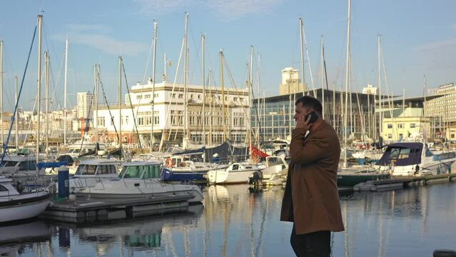 A male entrepreneur goes and calls on the phone in the port with expensive yachts, in sunny weather.