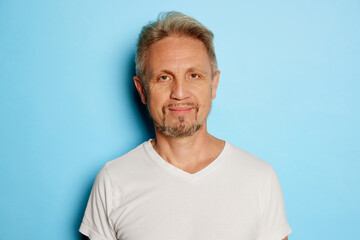 Obraz premium Portrait of mature man in white T-shirt posing, looking at camera isolated over blue studio background