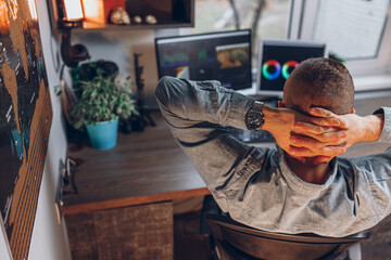 High angle back view of male video maker relaxing with hands behind head during video editing on computer monitors while working in home office 