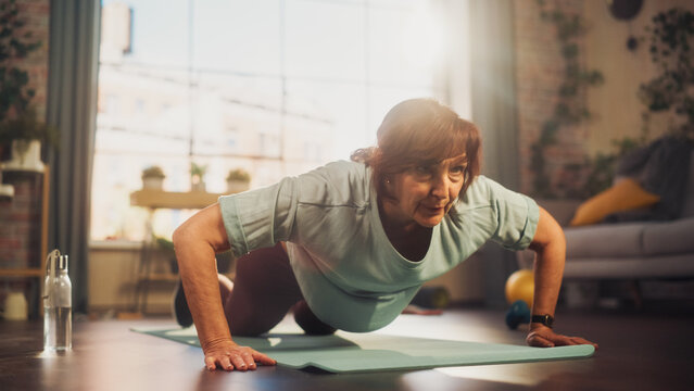 Happy Fit Senior Woman Training on a Yoga Mat, Doing Burpees Exercises During Morning Workout at Home in Sunny Apartment. Healthy Lifestyle, Fitness, Recreation and Retirement.