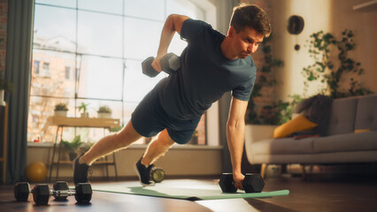 Fit Athletic Strong Young Man Performing Enduring Training in Plank Position, while Lifting Dumbbells During Morning Workout at Home in Sunny Apartment. Concept of Healthy Lifestyle and Fitness.