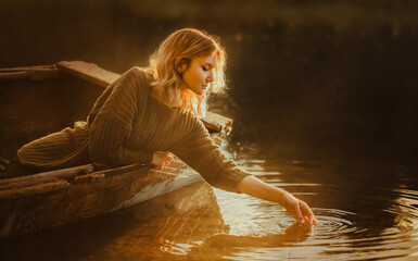 A beautiful blonde woman sits in an old fishing boat floating in the lake and touches the water...