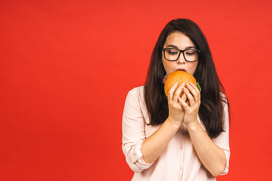 Portrait of young beautiful hungry woman eating burger. Isolated portrait of student with fast food over red background. Diet concept.