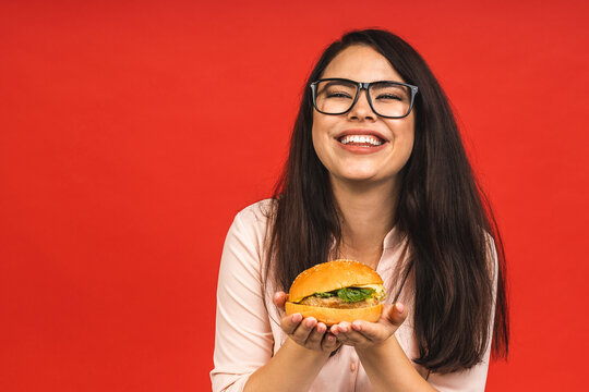 Portrait of young beautiful hungry woman eating burger. Isolated portrait of student with fast food over red background. Diet concept.