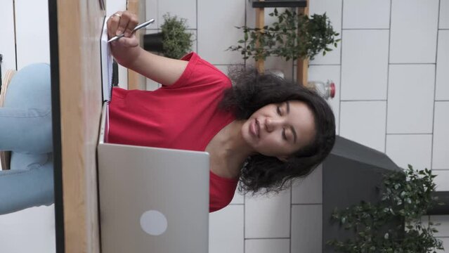 Young African American woman has video call using laptop for freelance work at home. Curly-haired lady in red T-shirt makes notes in paper notebook at wooden table