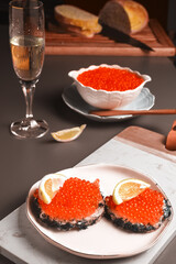 two sandwiches with red fish caviar served with a glass of champagne and a slice of lemon, in the background a porcelain white bowl filled with caviar with a teaspoon and sliced wheat bread