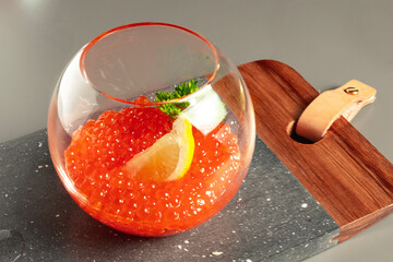 red caviar in a glass bowl with a spoon, served on a marble board with a slice of lemon and a sprig of parsley