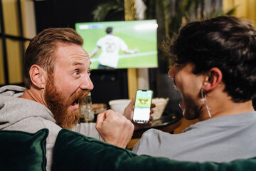 Two men watching football match and making bets at bookmaker's website in front of TV screen