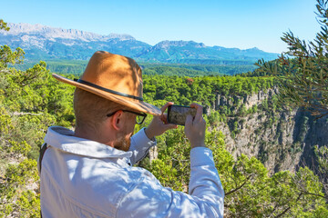 Caucasian male white shirt and brown hat takes pictures on smartphone of views on cliff near front...