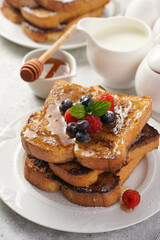 Several pieces of french toast - white wheat bread soaked in egg, milk and sugar, fried on a pan - stacked on white plate with fresh raspberries, blueberries, honey, mint on light grey background
