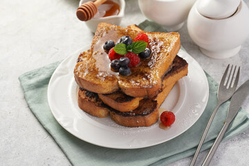 Several pieces of french toast - white wheat bread soaked in egg, milk and sugar, fried on a pan - stacked on white plate with fresh raspberries, blueberries, honey, mint on light grey background