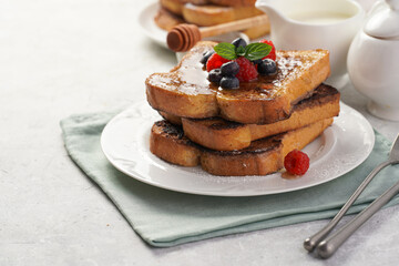 Several pieces of french toast - white wheat bread soaked in egg, milk and sugar, fried on a pan - stacked on white plate with fresh raspberries, blueberries, honey, mint on light grey background