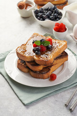 Several pieces of french toast - white wheat bread soaked in egg, milk and sugar, fried on a pan - stacked on white plate with fresh raspberries, blueberries, honey, mint on light grey background