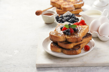 Several pieces of french toast - white wheat bread soaked in egg, milk and sugar, fried on a pan - stacked on white plate with fresh raspberries, blueberries, honey, mint on light grey background