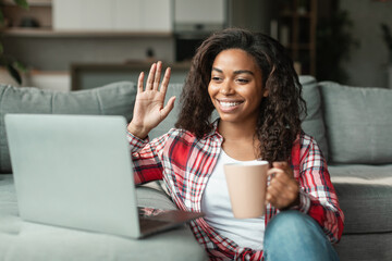 Cheerful pretty black young woman in casual with cup of hot drink waving her hand at laptop webcam