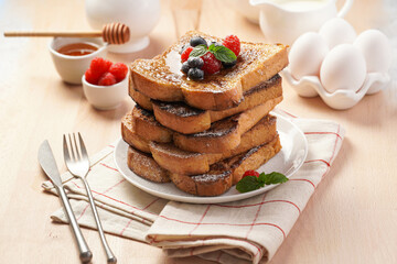 Several pieces of french toast - white wheat bread soaked in egg, milk and sugar, fried on a pan - stacked on white plate with fresh raspberries, blueberries, honey, mint on wooden background