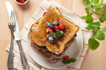 Several pieces of french toast - white wheat bread soaked in egg, milk and sugar, fried on a pan - stacked on white plate with fresh raspberries, blueberries, honey, mint on wooden background