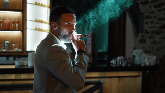 Handsome man holding cigar and looking at camera in dark luxury interior. Successful businessman smoking cigar